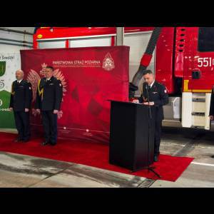 Firefighters in formal attire at an event, speaking at a podium in front of a fire truck and red backdrop.