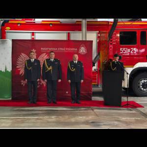 Three uniformed officials stand before a red backdrop, with a fire truck in the background during a formal event.
